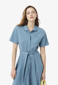 Vestito polo azzurro chiaro a maniche corte con bottoni, cintura in vita e un piccolo logo verde. Tessuto morbido con una vestibilità comoda.