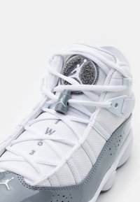 Vita och grå basketkor med texturerad mesh-överdel, glänsande lackläderdetaljer, logopatch och snörning.