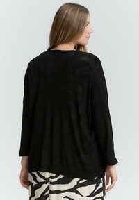 Černý lehký cardigan s dlouhými rukávy, uvolněný střih a mírně průhledná textura. Nositý přes vzorovanou krémovo-černou sukni.