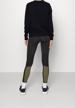 Persona vista desde atrás con una sudadera negra, leggings negro y verde oliva, calcetines blancos y zapatillas negras y blancas, de pie contra una pared blanca.