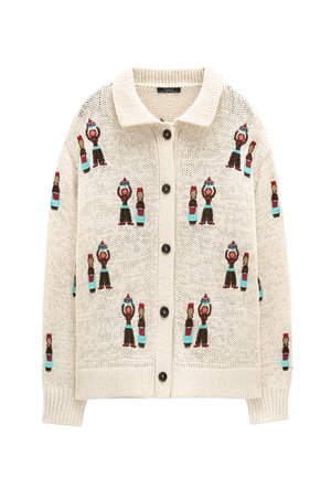 Beżowy dziergany sweter na guziki, z powtarzającym się wzorem małych postaci w kapeluszach i niebieskich spodniach obok butelek z czerwonymi nakrętkami.