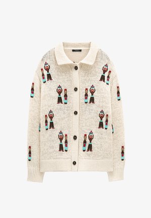 Beżowy dziergany sweter na guziki, z powtarzającym się wzorem małych postaci w kapeluszach i niebieskich spodniach obok butelek z czerwonymi nakrętkami.