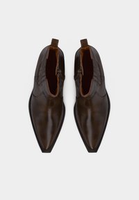 Ζευγάρι γυαλισμένων σκούρων καφέ δερμάτινων μποτάκια αστραγάλου με μυτερά μύτα και θηλιά έλξης, από πάνω, σε λευκό φόντο.