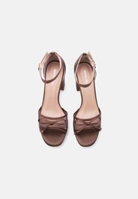 Καφέ σανδάλια με τακούνι και σατέν φινίρισμα, ανοιχτά δάχτυλα και διάφανοι σταυρωτοί ιμάντες. Διαθέτουν λουράκι στον αστράγαλο με μικρή αγκράφα.