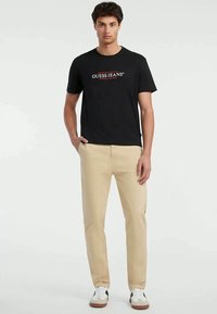 Νεαρός άνδρας φοράει μαύρο t-shirt με λογότυπο Guess, μπεζ παντελόνι και λευκά αθλητικά παπούτσια, στέκεται με το ένα χέρι στην τσέπη.