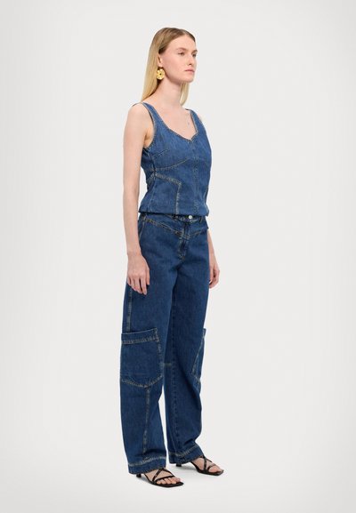 Farmermellény nélküli, középkék denim felső és széles lábú nadrág. Kiemelkedő varrásokkal és zsebfoltokkal rendelkezik. A modellen fekete pántos szandál van.