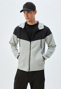 Graue und schwarze Zip-Jacke mit Kapuze, langen Ärmeln, Fronttaschen und einem kontrastierenden Farbblockdesign. Schwarze Baseballmütze.