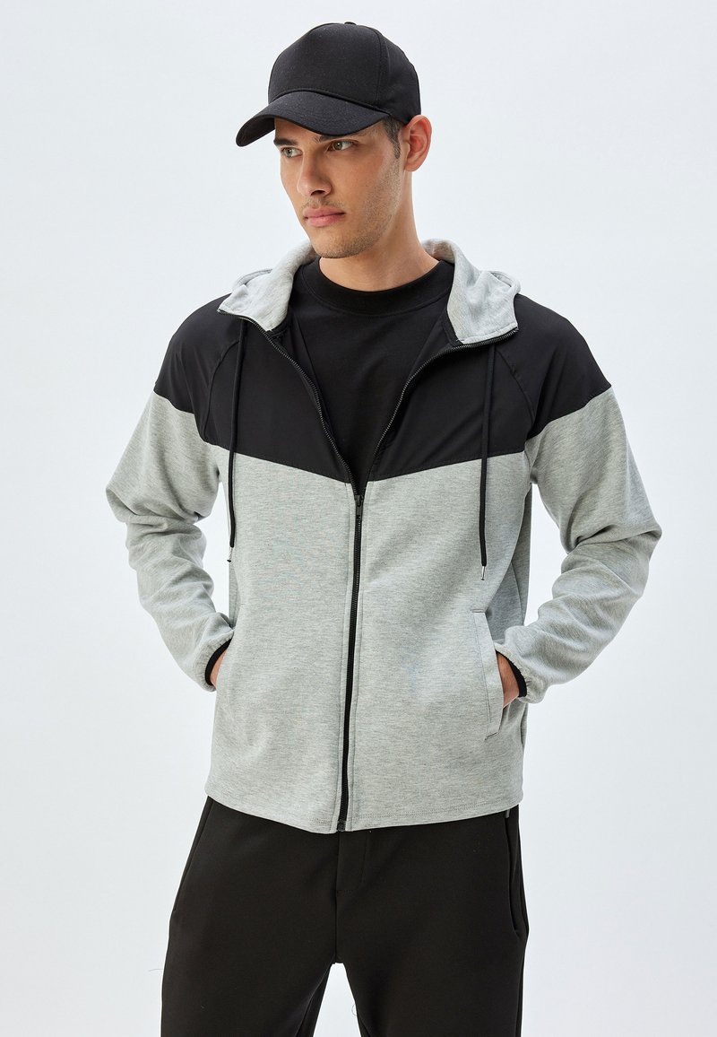 Grå och svart zip-up jacka med huva, med långa ärmar, framfickor och en kontrasterande färgblockdesign. Svart basebollkeps.