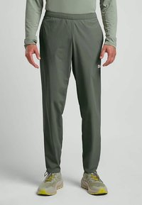 Man in olijfgroene taps toelopende sweatpants met elastische tailleband en lichtgrijze hardloopschoenen met gele zolen en veters, rechtopstaand.