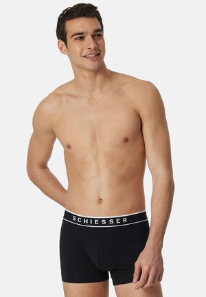 Man zonder shirt die zwarte Schiesser boxershorts draagt, staand met één hand achter zijn rug en glimlachend, tegen een effen achtergrond.