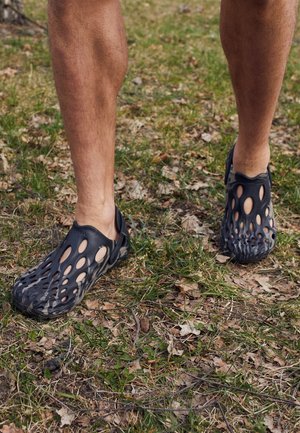 Personne portant des chaussures slip-on noires à motif de trous, se tenant sur de l'herbe et des feuilles sèches à l'extérieur.