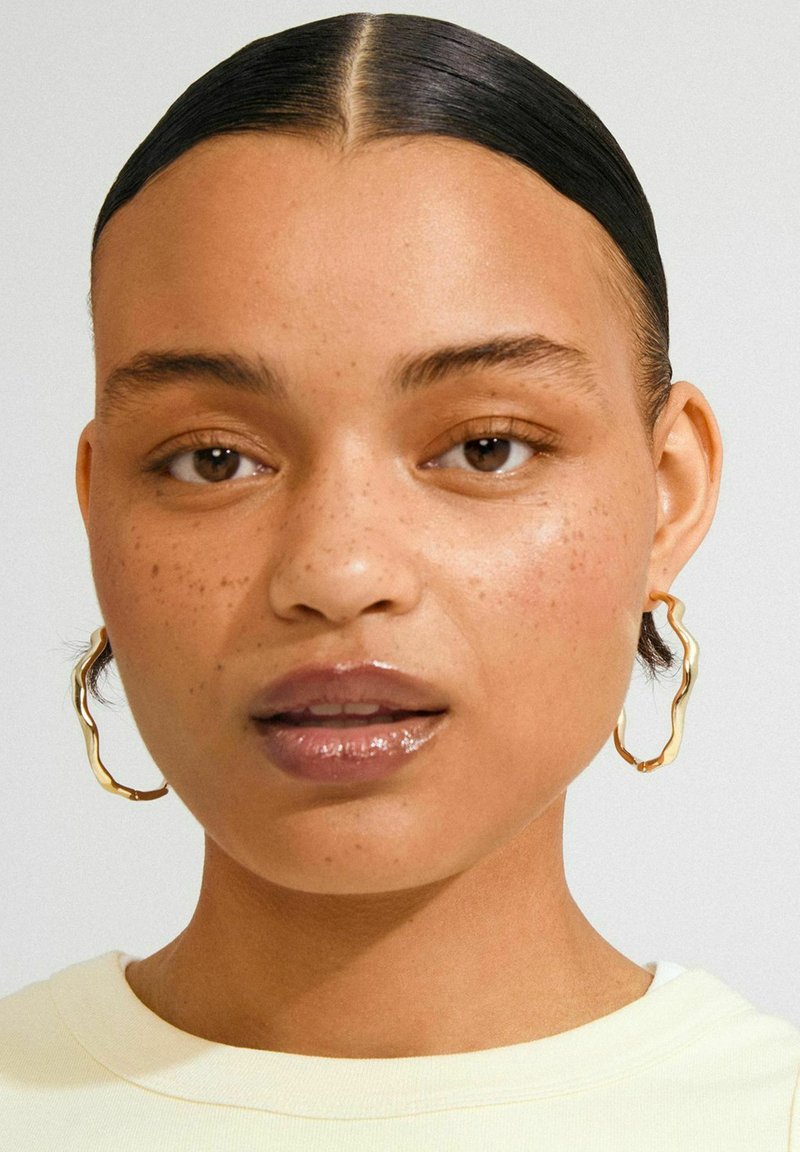 Donna con capelli lisci e raccolti, lentiggini leggere, labbra lucide, che indossa grandi orecchini a cerchio in oro di forma irregolare e una maglietta gialla pallida.