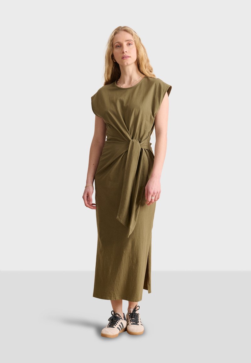 Abito verde oliva, lungo fino al ginocchio, con fiocco frontale, maniche corte a campana e uno spacco laterale, abbinato a sneakers beige con lacci neri.