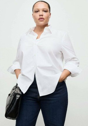 Donna adulta che indossa una camicia bianca abbottonata e jeans scuri con le mani nelle tasche, tenendo una borsa nera contro uno sfondo semplice.