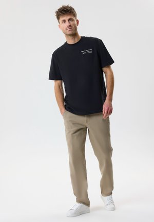 Lühikeste juustega mees, kes kannab musta trükitud t-särki, beeže pükse ja valgeid vabaajajalatsid, seisab käed taskutes valgel taustal.