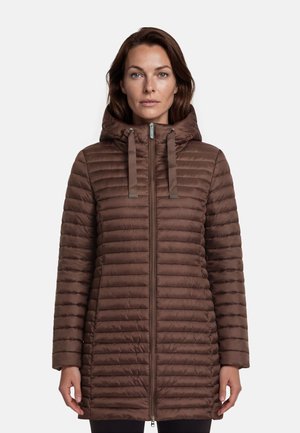 Femme portant une longue veste matelassée marron avec capuche et zipper frontal, debout devant un fond clair uni.