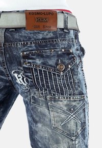 Jeans in denim con un lavaggio blu sbiadito, decorati con cuciture, numerose tasche e accenti metallici. Presentano un'etichetta in pelle sulla vita.