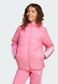 Junge Frau mit lockigem Haar lächelt, trägt eine rosa Daunenjacke und passende Hose mit weißen Streifen und einem Adidas-Logo.