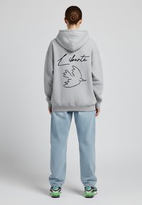 Felpa grigia con un contorno nero di una colomba e la parola "Liberté" sulla schiena; abbinata a pantaloni azzurri e sneakers.