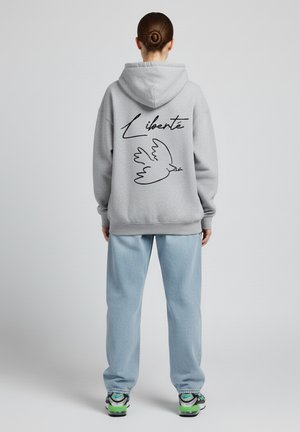 Grijze hoodie met een zwarte omtrek van een duif en het woord "Liberté" op de achterkant; gecombineerd met lichtblauwe broek en sneakers.