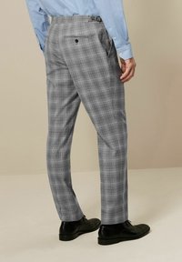Un pantalon à carreaux gris présente une coupe ajustée, avec une poche arrière simple et des passants de ceinture discrets, associé à des chaussures de ville noires.