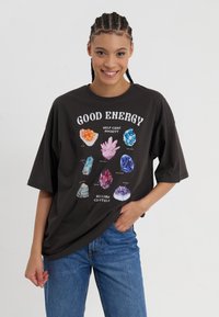 T-shirt noir oversize avec un graphique coloré de divers cristaux accompagné du texte "BONNE ÉNERGIE". Le matériau semble doux et a une coupe décontractée.