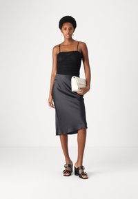 Melna caurspīdīga siksniņu tops, kas apvienots ar gludu, tumši pelēku satīna svārku. Modelis tur baltu blakus somu un valkā melnus papēžu sandales.