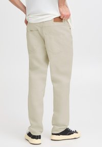 Pantalones beige claro de corte recto, con dos bolsillos traseros, costuras sutiles y un parche de cuero marrón en la cintura.