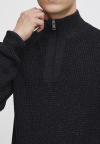Schwarzer gestrickter Pullover mit hohem Kragen und einem halben Reißverschluss. Die Struktur wirkt grob und verstärkt die Wärme und den Komfort.