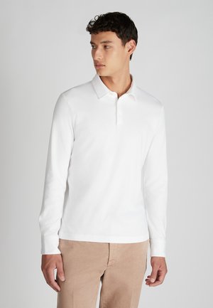 Jongeman in een witte poloshirt met lange mouwen en beige broek, kijkend naar links tegen een effen lichte achtergrond.