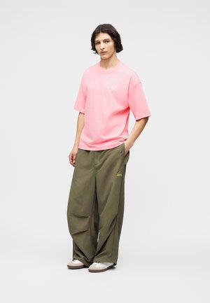 Jonge persoon staand met hand in de zak, gekleed in een losse roze korte mouwen shirt, wijde olijfgroene Adidas broek en witte sneakers.