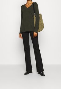 Femme portant un pull col V vert foncé, pantalon noir évasé, bottes noires à talons et un sac à bandoulière en suède olive.