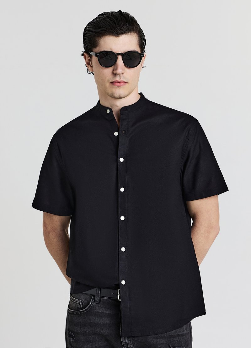 Chemise noire à manches courtes avec col mandarin, dotée d'une fermeture à boutons sur le devant et d'une texture lisse. Le modèle porte des lunettes de soleil rondes noires.