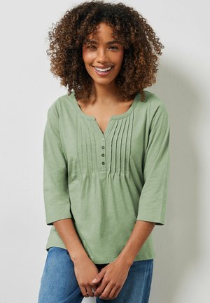 Femme souriante aux cheveux bouclés portant un haut vert clair à manches trois-quarts avec des boutons sur le devant et des plis, debout devant un mur uni.