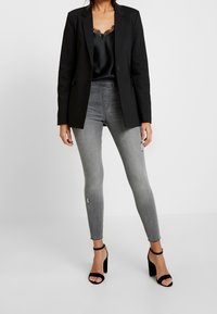 Blazer nero su una canotta di satin nera con bordo in pizzo, abbinato a jeans skinny grigi strappati e sandali neri con tacco alto.