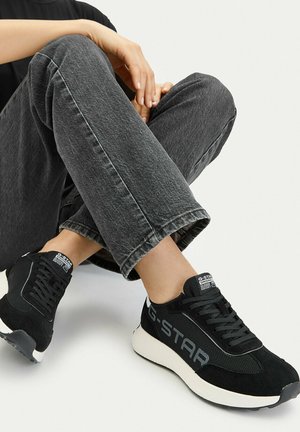 Osoba sedící s překříženýma nohama, obutá v černých teniskách G-Star a tmavě šedých džínách, na jednobarevném bílém pozadí.