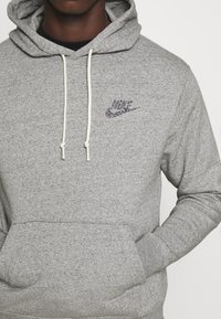 Šedá fleecová mikina s kapucí na stahování, přední kapsou a vícebarevným logem Nike na hrudi. Strukturovaná tkanina s mírnými tečkami.