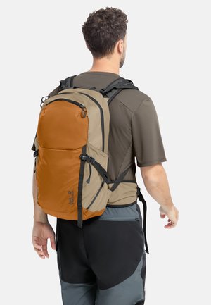 Homme portant un sac à dos de randonnée bicolore marron et beige avec plusieurs sangles, vêtu d'une chemise marron et d'un pantalon noir-gris, dos tourné.