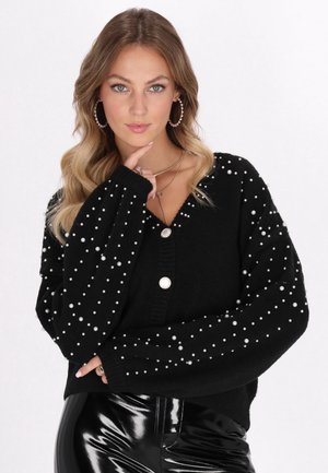 Schwarze Strickjacke mit V-Ausschnitt, verziert mit weißen Perlen. Sie verfügt über eine Knopfschließe und weite Ärmel, kombiniert mit glänzenden schwarzen Hosen.