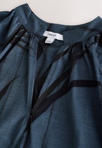 Blå stofbluse med rund halsudskæring, plisseret design og abstrakt sort mønster. Mærket siger "NEXT." Glat tekstur og let vægt.
