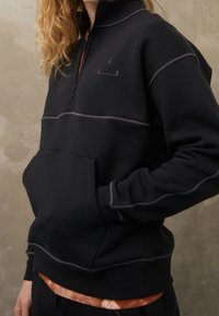 Sudadera negra con cremallera, costuras de acento grises, bolsillo frontal y logo sutil. El material parece suave y texturizado.