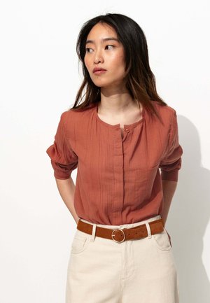 Blouse couleur rouille avec un col rond, fermeture à boutons et manches courtes bouffantes, assortie à un pantalon beige clair et une ceinture marron.