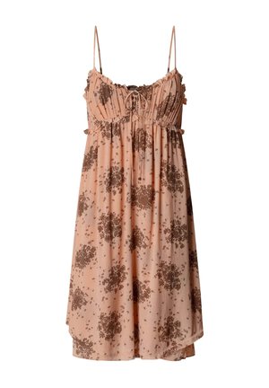 Jeune femme aux longs cheveux blonds portant une robe rose sans manches avec des imprimés floraux marron, regardant sur le côté sur un fond uni.