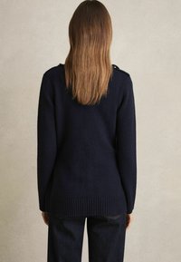 Pulover din tricot bleu marin cu tiv ridicat, nasturi pe umeri și mâneci lungi. Textură moale, croială ușor lejeră, perfect pentru a fi purtat în straturi.