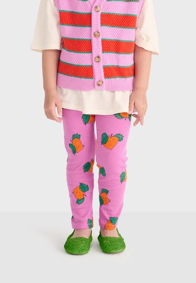 Enfant portant un gilet en tricot rayé rose et rouge, des leggings roses avec des motifs de fruits orange, et des chaussures en tricot vertes, debout devant un fond uni.