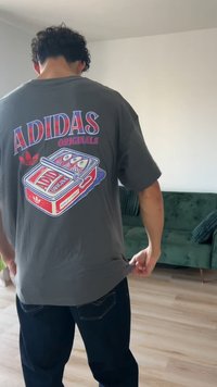 Γκρι βαμβακερό μπλουζάκι με τυπωμένο κόκκινο και μπλε κείμενο "ADIDAS ORIGINALS" και γραφικό σχέδιο με κονσέρβα, φορεμένο με σκούρο μπλε τζιν.