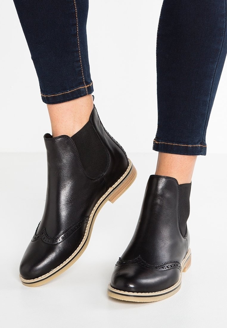 Bottes chevilles en cuir noir avec détails brogue et semelles beiges portées avec un jean skinny bleu foncé retroussé sur fond clair.