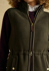 Olivovo zelená fleecová vesta s vysokým golierom, plným zipsom, hnedými akcentmi a bočnými vreckami, s detailom stiahnutého pásu.