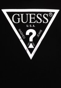 Schwarzes T-Shirt mit einem reflektierenden weißen Dreieck-Logo, das den Text "GUESS" und "U.S.A." zeigt, sowie den Etiketten "WASHED" und "JEANS" im Inneren.