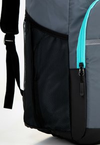 Sac à dos gris avec une poche latérale en mailles noires, une fermeture éclair turquoise et un tissu texturé. Comprend des bretelles rembourrées pour plus de confort.